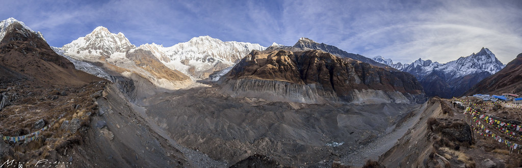 Himalayan glacial valley with snow-covered peaks, rocky slopes, prayer flags, and blue-roofed mountain settlement.
