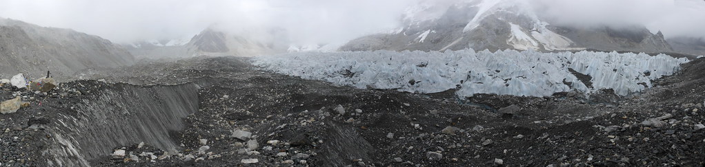Jagged bluish-white glacier with deep crevasses, surrounded by rocky moraine and misty snow-covered mountains.