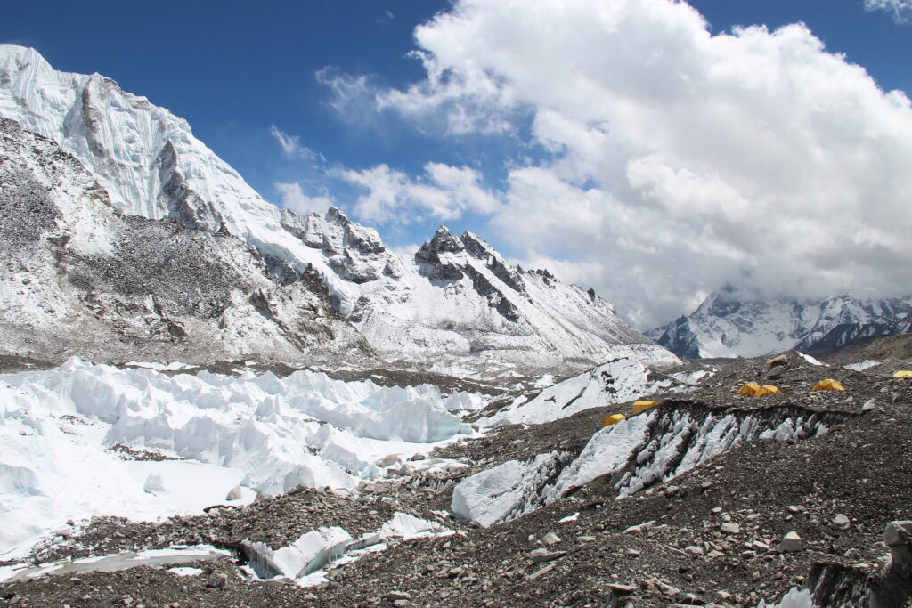 Snow-covered Himalayan peak with icy slopes, rocky valley, trekkers, and tents at high-altitude campsite.