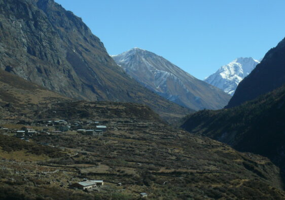 A rugged Himalayan valley cradles a small stone village beneath towering snow-capped peaks, where human life clings to terraces carved into the earth under a vast, clear sky.