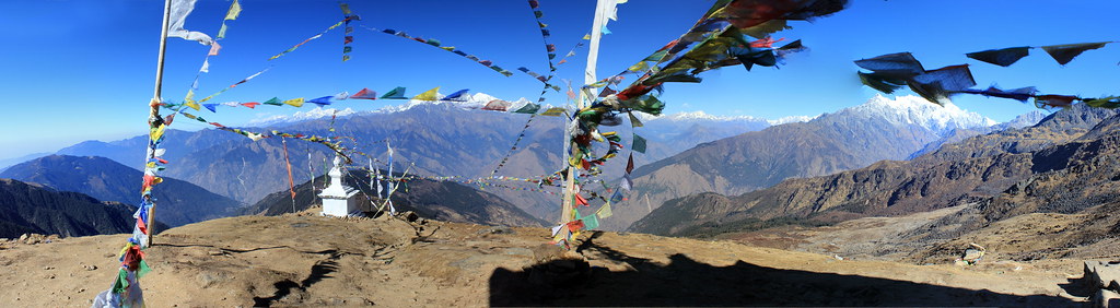 White Himalayan stupa with colorful prayer flags on rocky plateau, snowy peaks in background.