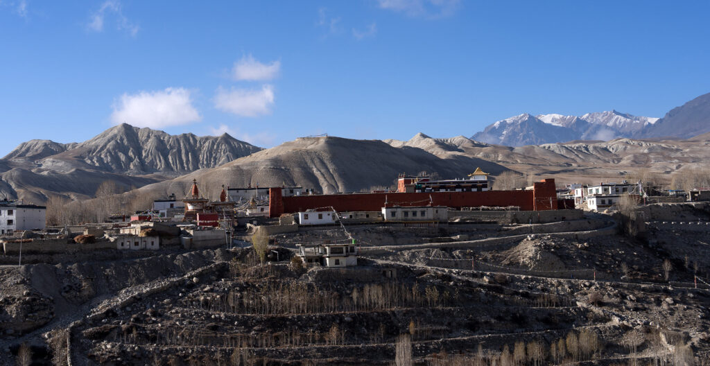 Himalayan village with red-walled monastery, golden roofs, terraced slopes, and snowy peaks in background.
