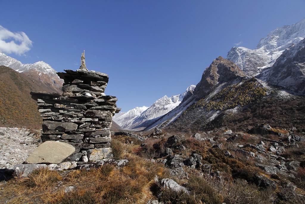 Stone cairn or stupa on rocky Himalayan slope with sparse vegetation and snowy peaks in background.
