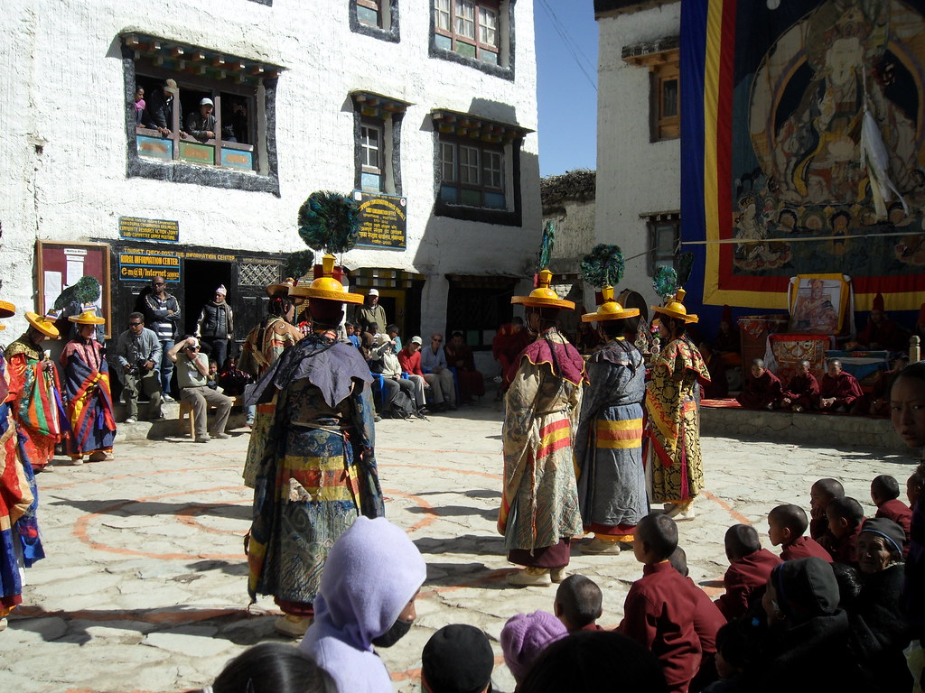 Tibetan Buddhist dance performance in courtyard, with performers in colorful robes and tall yellow hats, watched by monks and spectators.