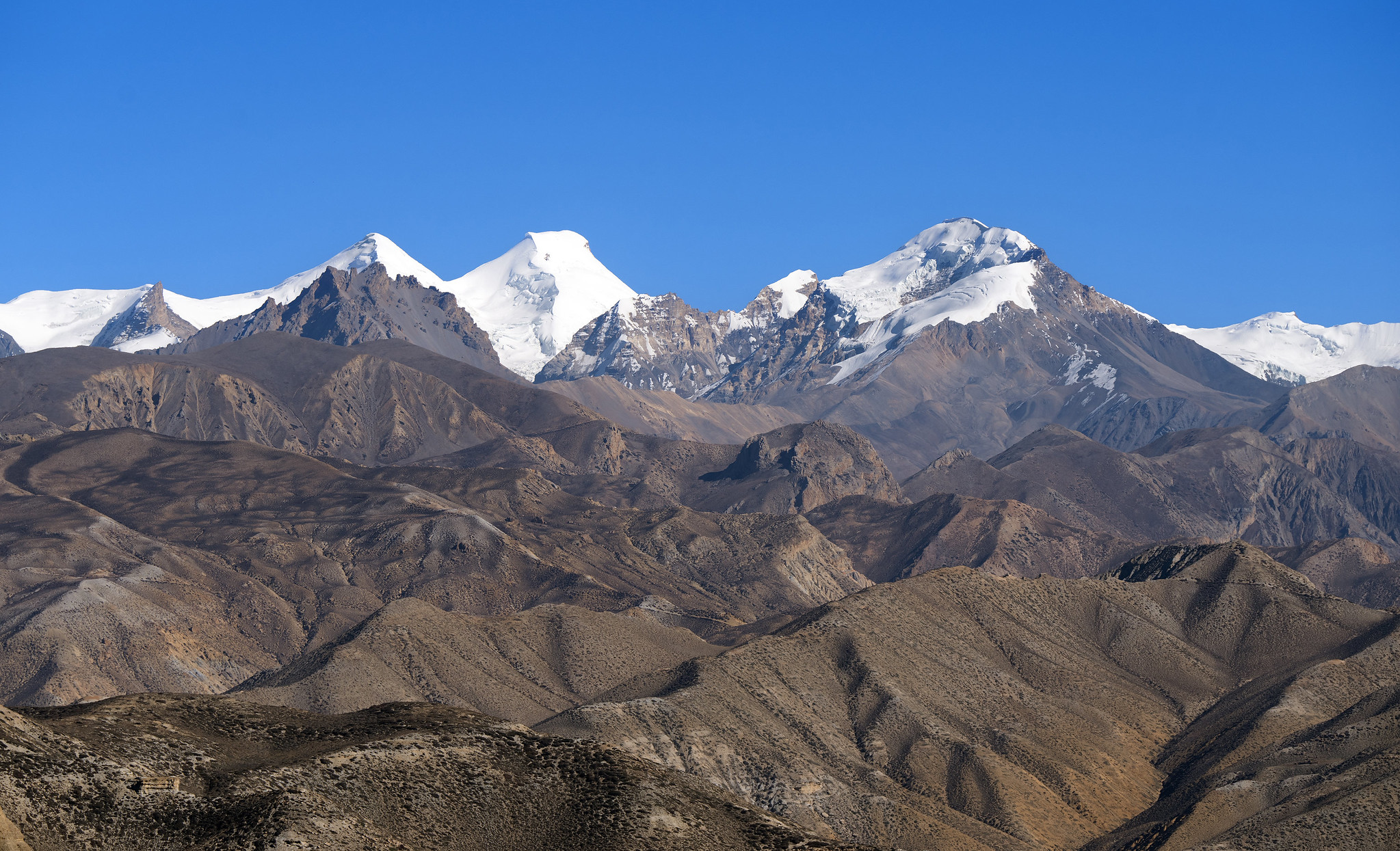 Rugged brown mountain ridges leading to towering snow-capped peaks under a deep blue sky.