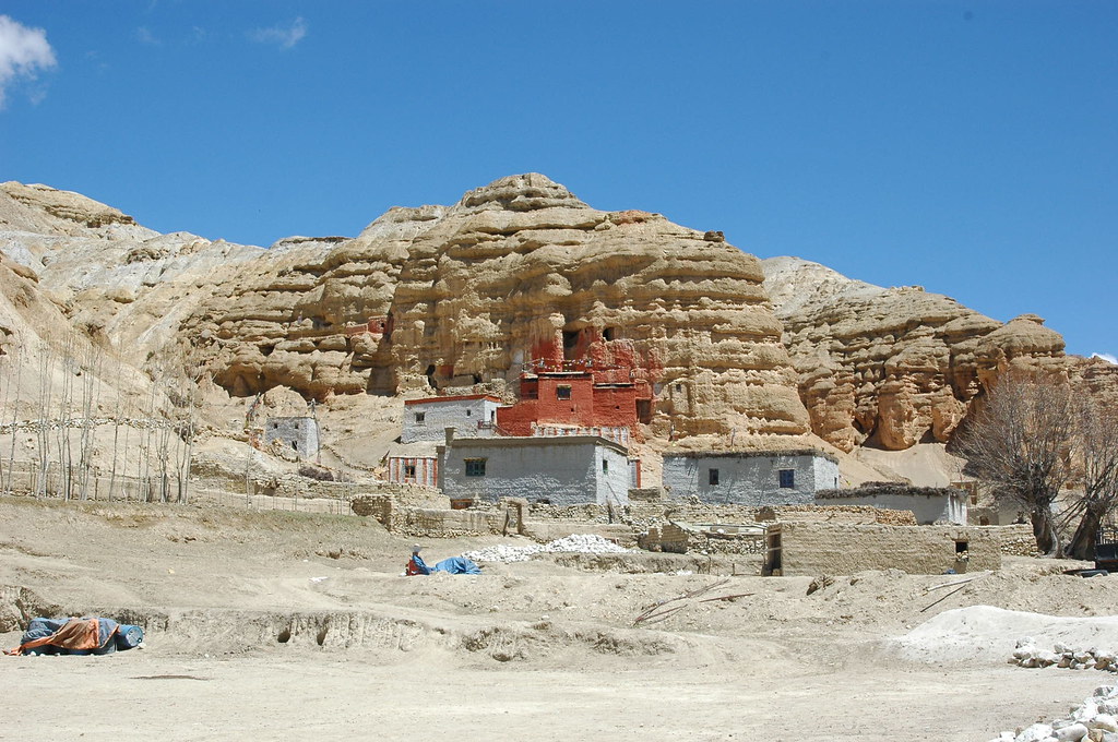 Cluster of traditional Himalayan buildings at the base of layered sandstone cliffs, with vivid red monastery.