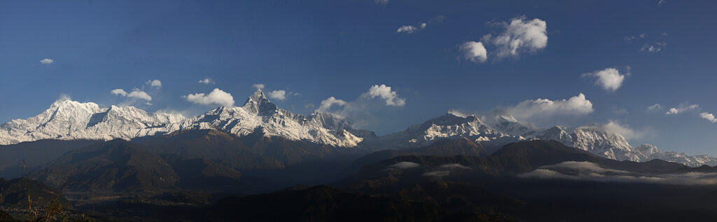 Panoramic Himalayan mountain range with central snow-covered peak, forested lower slopes, and clear blue sky.