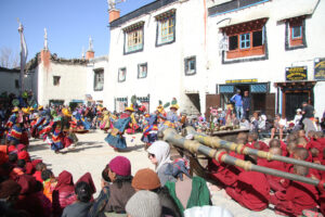 Traditional Buddhist dance performance in a monastery courtyard, with performers in multicolored robes and tall yellow hats adorned with green feathers. A large tapestry of a deity hangs on the wall, monks in maroon robes and other spectators watch from the ground and windows above.
