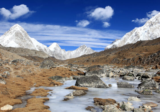 High-altitude Himalayan landscape with snow-covered peaks, rocky tundra, and a partially frozen stream under a blue sky.