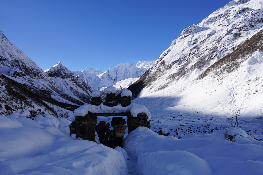 Trekkers passing through a snow-covered stone archway in a Himalayan valley beneath rugged peaks