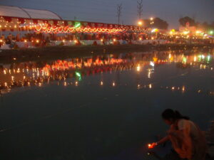 Evening river side chhath festival with lights and people placing lamps on water.