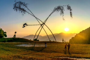 Person swinging high on a traditional bamboo swing at sunset in a grassy field with hills.