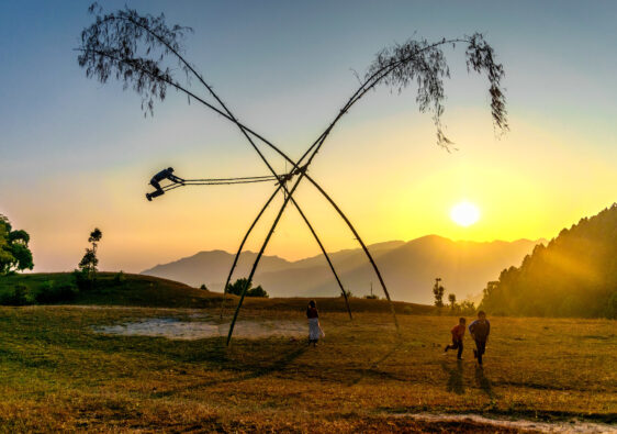 Person swinging high on a traditional bamboo swing at sunset in a grassy field with hills.