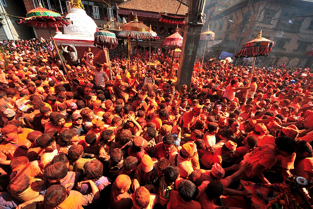 Crowd covered in orange and red powder celebrating a vibrant festival in a Nepalese square with ornate umbrellas and stupa.