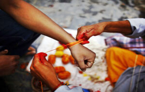 Hands tying yellow-orange thread on wrist during ritual with offerings nearby.