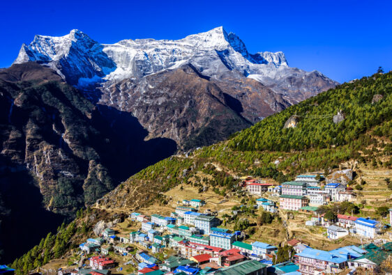 “Colorful Himalayan village on mountain slope with snowy peaks in background”